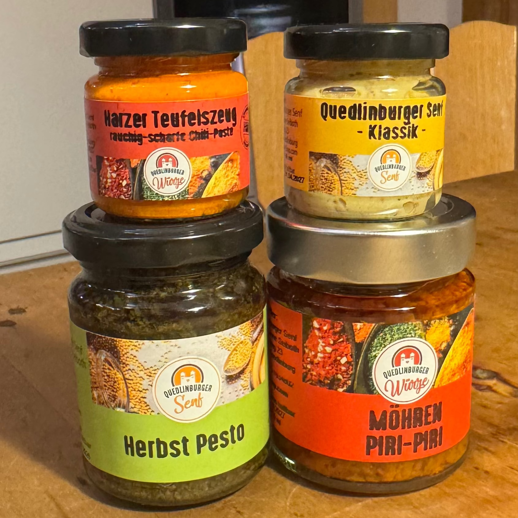 Vier kleine Gläser (Klassik-Senf, Möhren-Piri-Piri, Kräuterpesto, Teufelszeug) als veganes Probier-Set, arrangiert in natürlicher Küchen- oder Holzoptik – mild bis scharf.