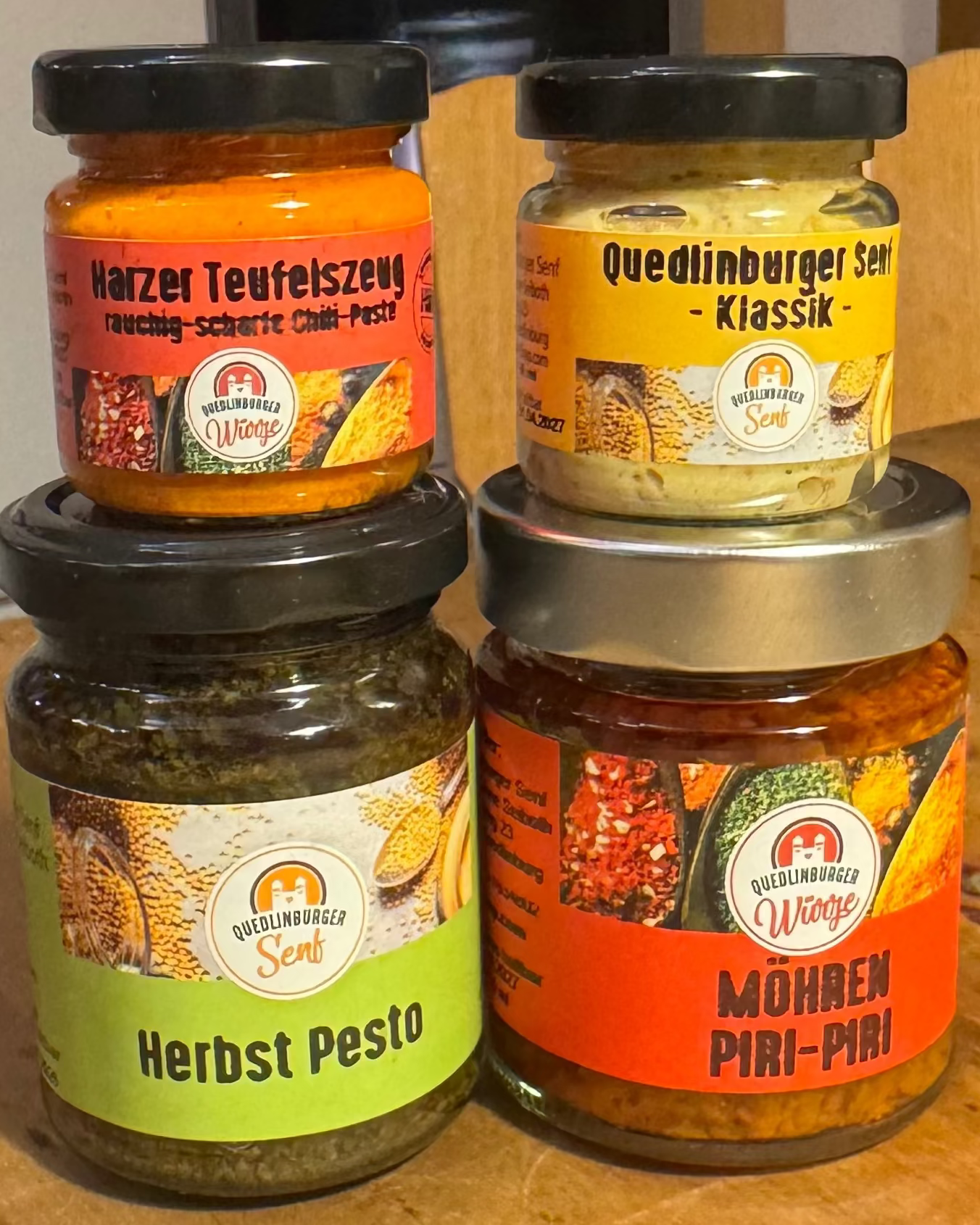 Vier kleine Gläser (Klassik-Senf, Möhren-Piri-Piri, Kräuterpesto, Teufelszeug) als veganes Probier-Set, arrangiert in natürlicher Küchen- oder Holzoptik – mild bis scharf.