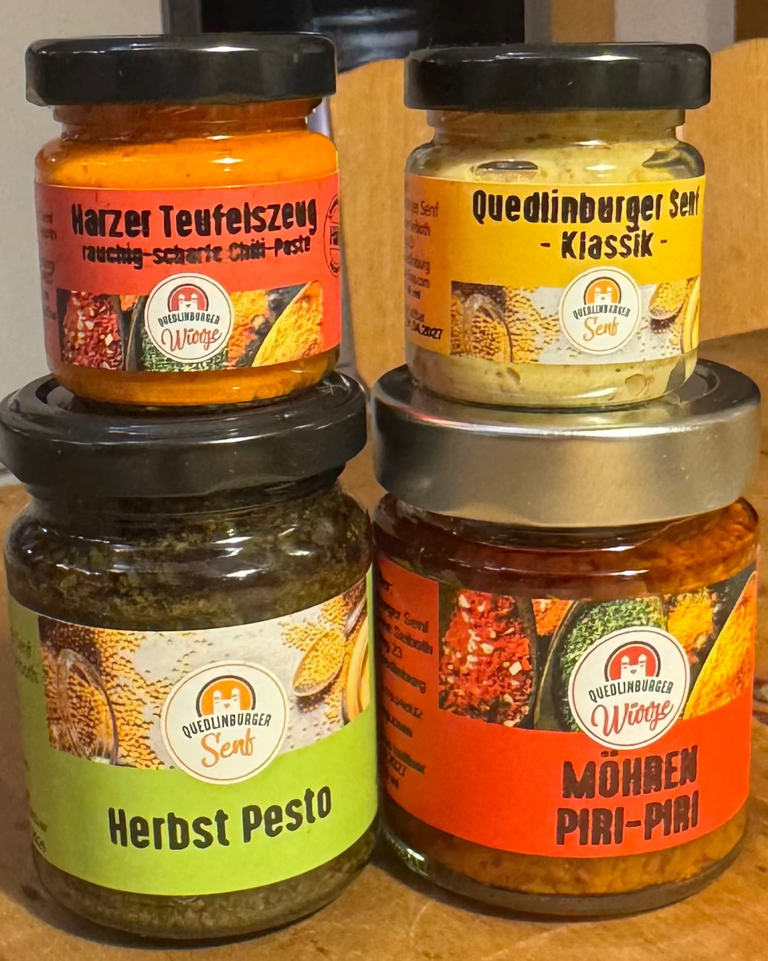 Vier kleine Gläser (Klassik-Senf, Möhren-Piri-Piri, Kräuterpesto, Teufelszeug) als veganes Probier-Set, arrangiert in natürlicher Küchen- oder Holzoptik – mild bis scharf.