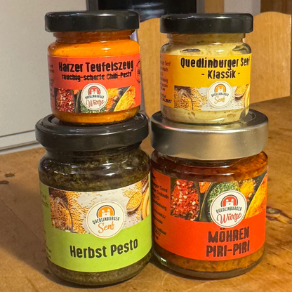 Vier kleine Gläser (Klassik-Senf, Möhren-Piri-Piri, Kräuterpesto, Teufelszeug) als veganes Probier-Set, arrangiert in natürlicher Küchen- oder Holzoptik – mild bis scharf.